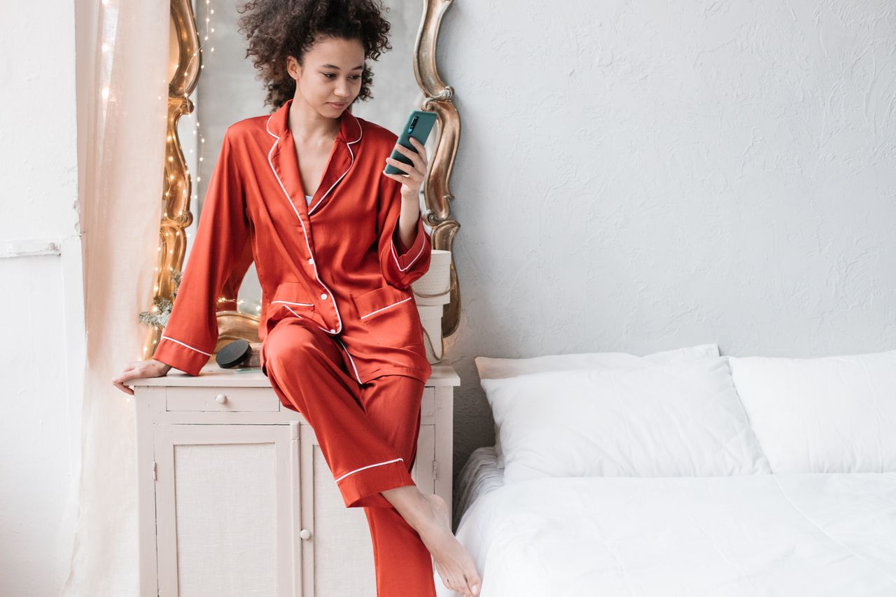 A woman in red pajamas sitting comfortably using a smartphone in a cozy bedroom setting.