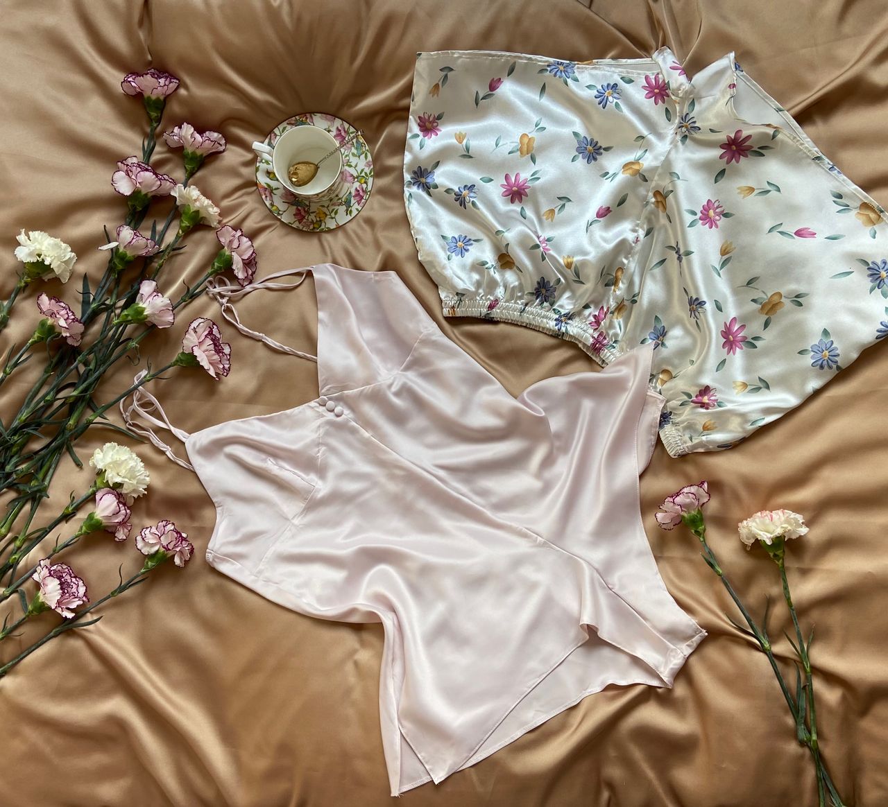 Elegant silk nightwear with colorful flowers on green stems placed on creased textile with ceramic cup in light room at home
