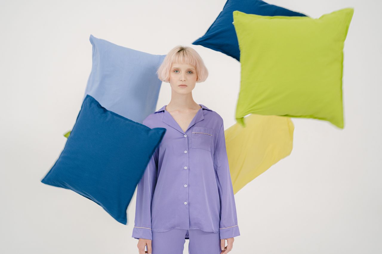 A woman in purple sleepwear surrounded by vibrant floating pillows on a white background.